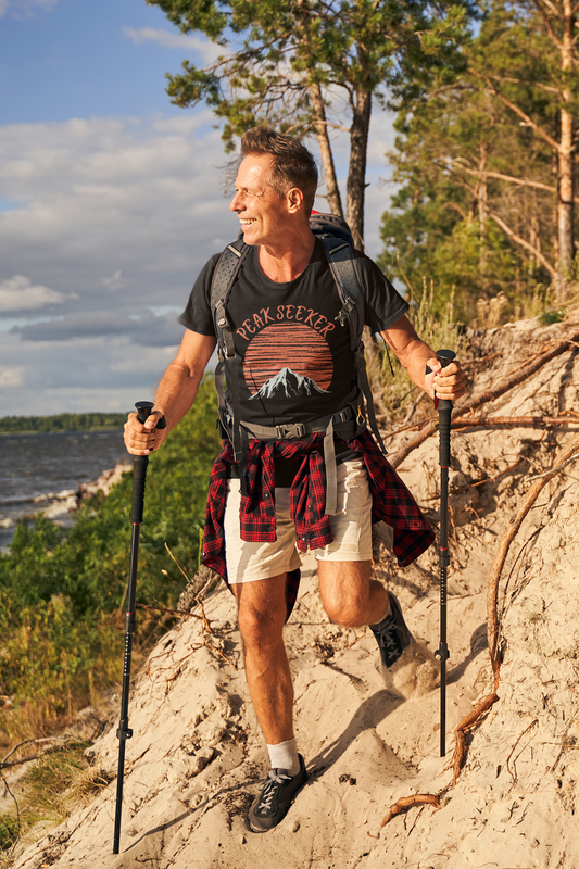 Man in shorts climbing up a mountain wearing black Peak Seeker T-Shirt with Peak Seaker in orange fancy type in an arc over an orange sunrise, moonrise or sunset with a mountain below silloetting the son.