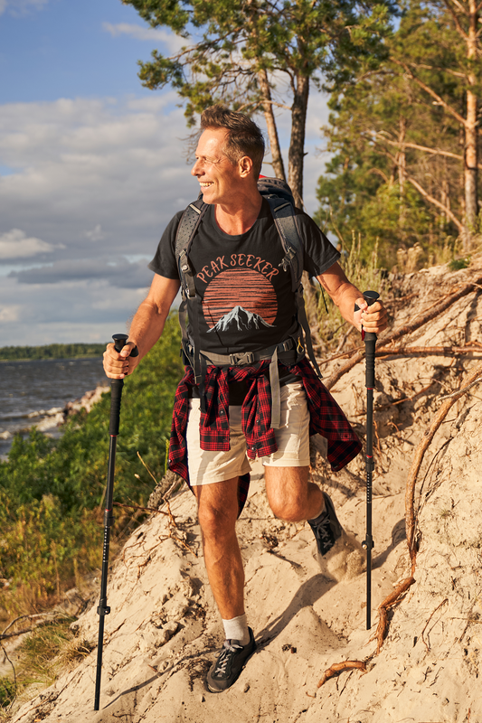 Man in shorts climbing up a mountain wearing black Peak Seeker T-Shirt with Peak Seaker in orange fancy type in an arc over an orange sunrise, moonrise or sunset with a mountain below silloetting the son. 