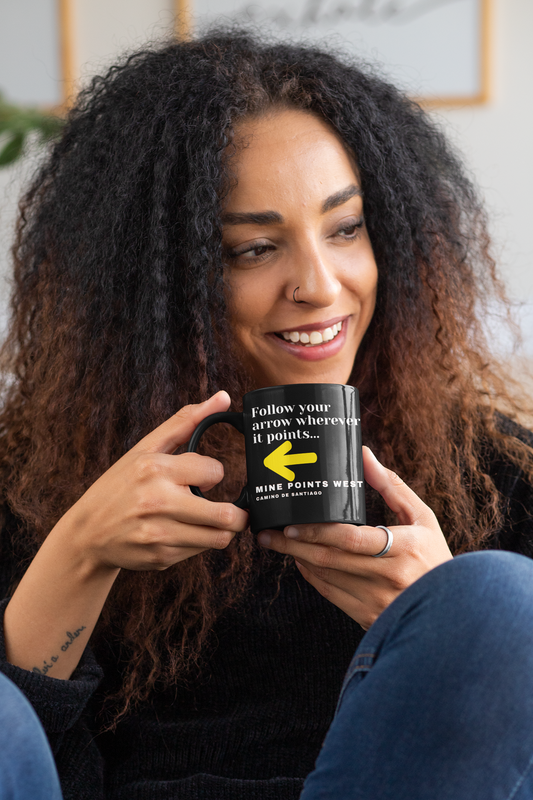 Camino Frances - A beautiful young woman wearing a dark shirt and blue jeans is sitting and is holding a black ceramic mug with  a slogan that says in white letter , Follow your arrow wherever it points . . . With a yellow arrow below it then beow that the words, Mine Points West and under that Camino de Santiago.