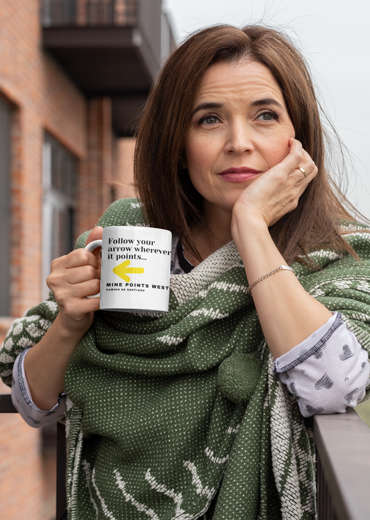 Camino Frances - A middle-aged woman dressed in a print shirt and a green and beige shawl is tanding on a patio with her left elbow on a railing and her left palm on the side of her cheek with her chin in her palm. In her right hand she is holding a white ceramic mug. This white ceramic mug has a slogan in black letters that says, Follow your arrow wherever it points . . . With a yellow arrow below it then below that the words, Mine Points West and under that Camino de Santiago.