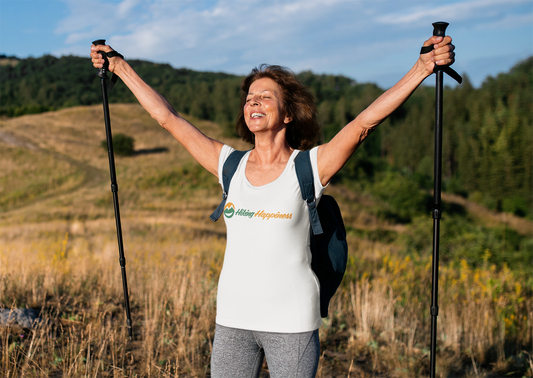 A middle aged lady is overjoyed and smiling with both arms raised with her trekking poles in both hands and a smile on her face. She is wearing a white cotton t-shirt with the Hiking Happiness smiling logo and the Words Hiking Happiness at chest level. She has her back pack on and behind her is a field surrounded with green trees. 