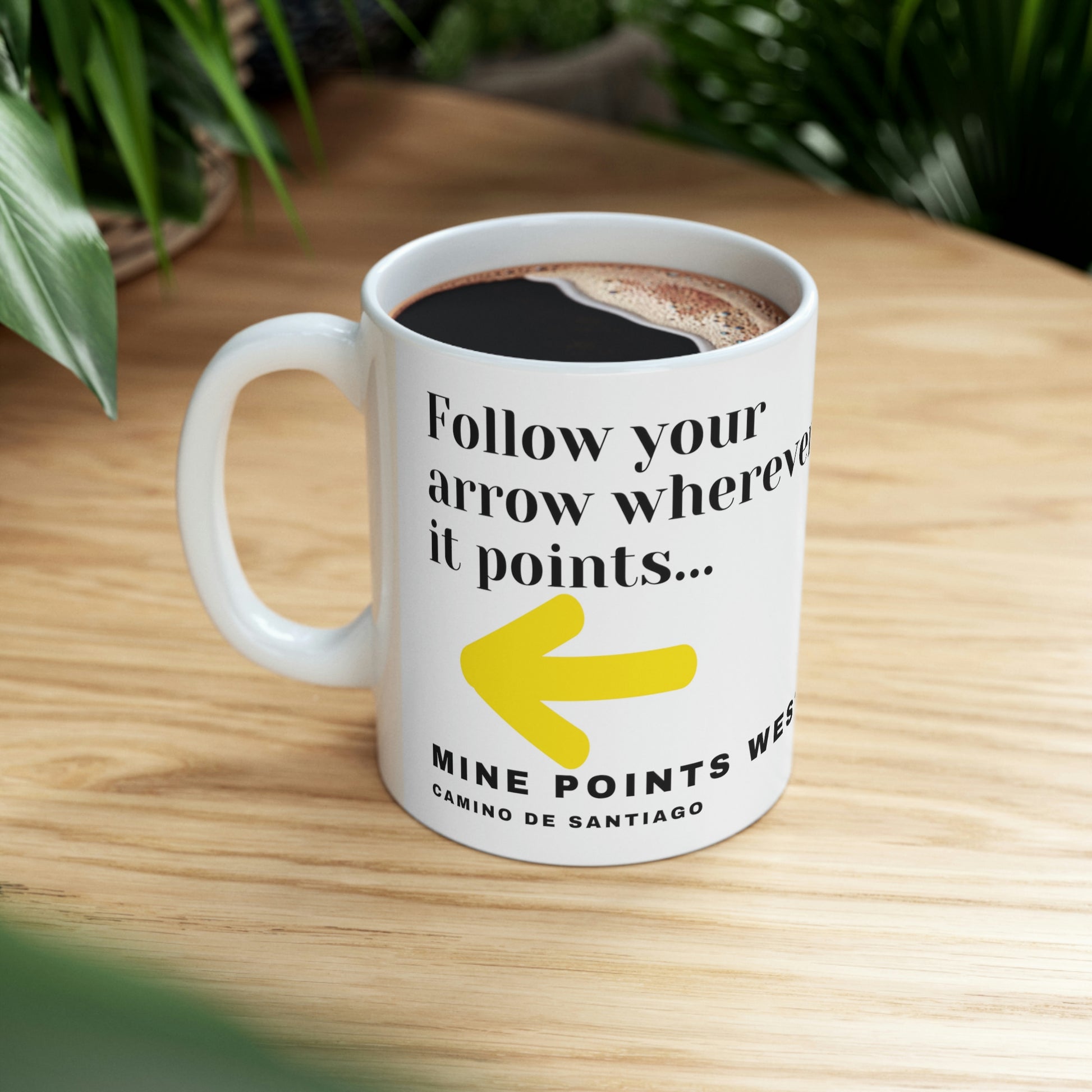 Camino Frances - This white ceramic mug has a slogan in clack letters that says, Follow your arrow wherever it points . . . With a yellow arrow below it then below that the words, Mine Points West and under that Camino de Santiago. The Mug is filled with hot coffe and is on a woodaen table with green plants in the background.